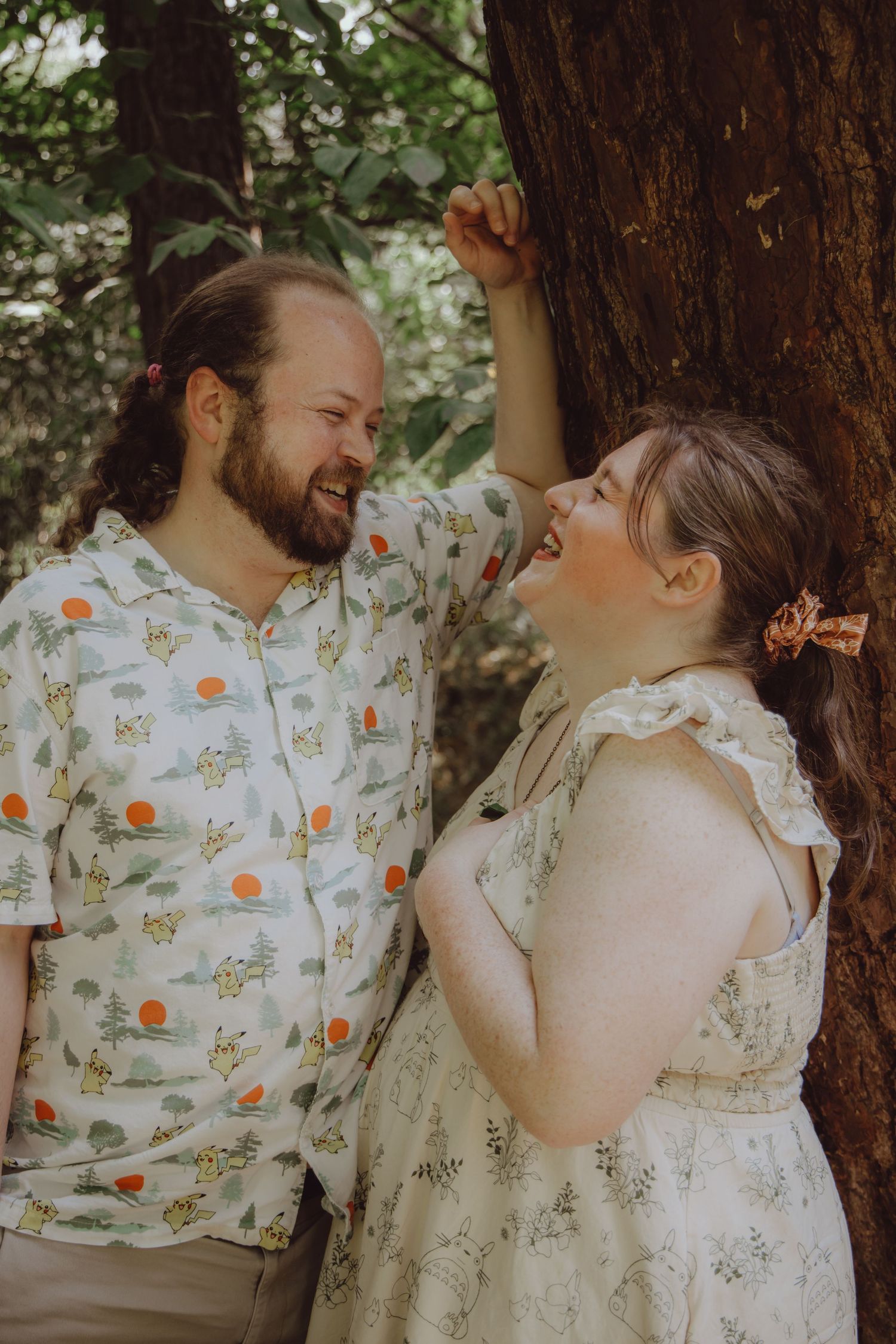 A couple laughs together leaning against a tree, man in Pikachu shirt, woman in floral dress outdoors.