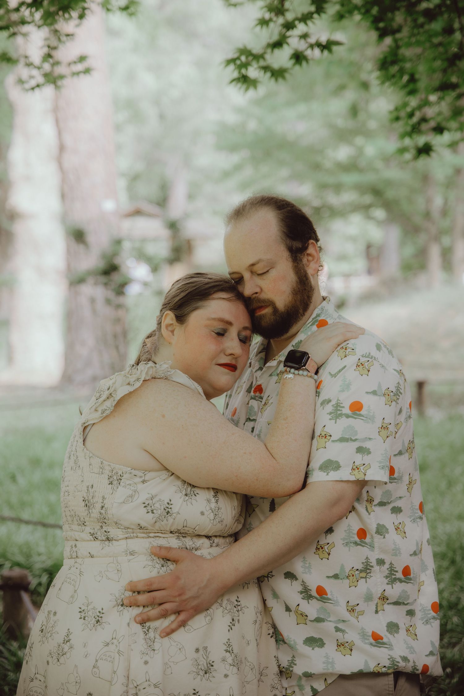 A couple embraces tenderly in a lush green park, the woman in a floral dress leaning into the bearded man holding her close.