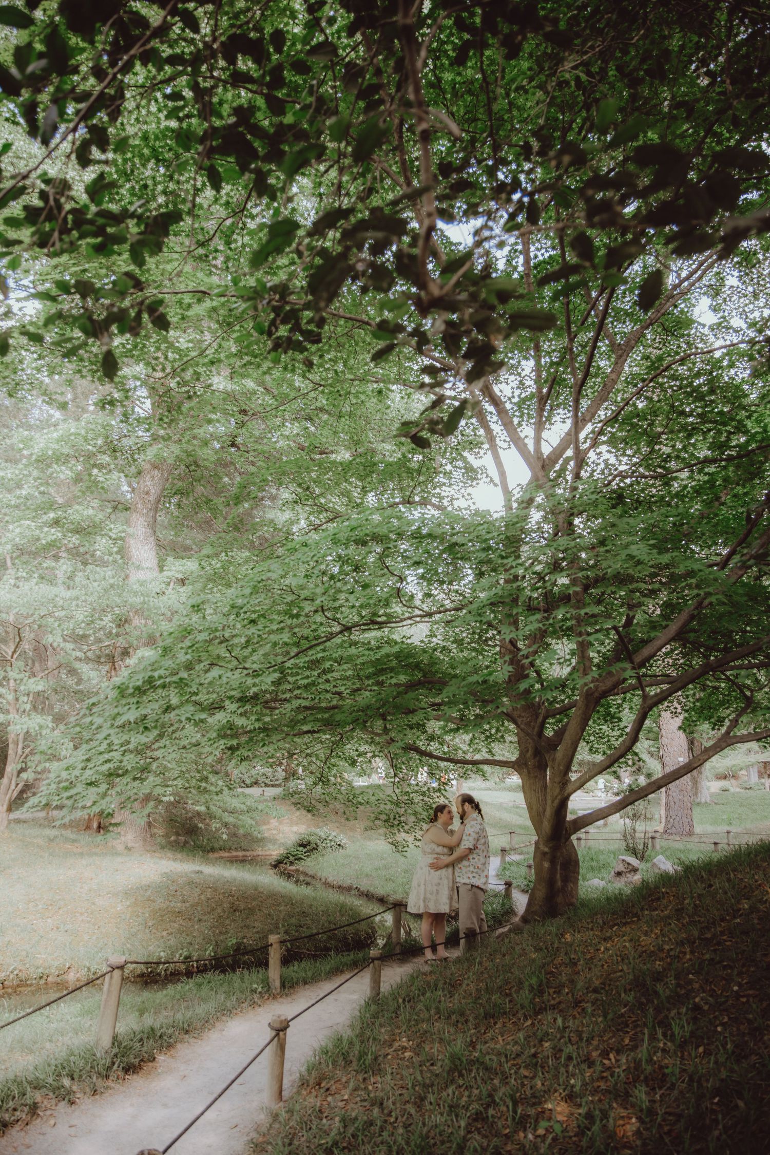 Couple embracing under lush green trees along a serene garden path with wooden railings.