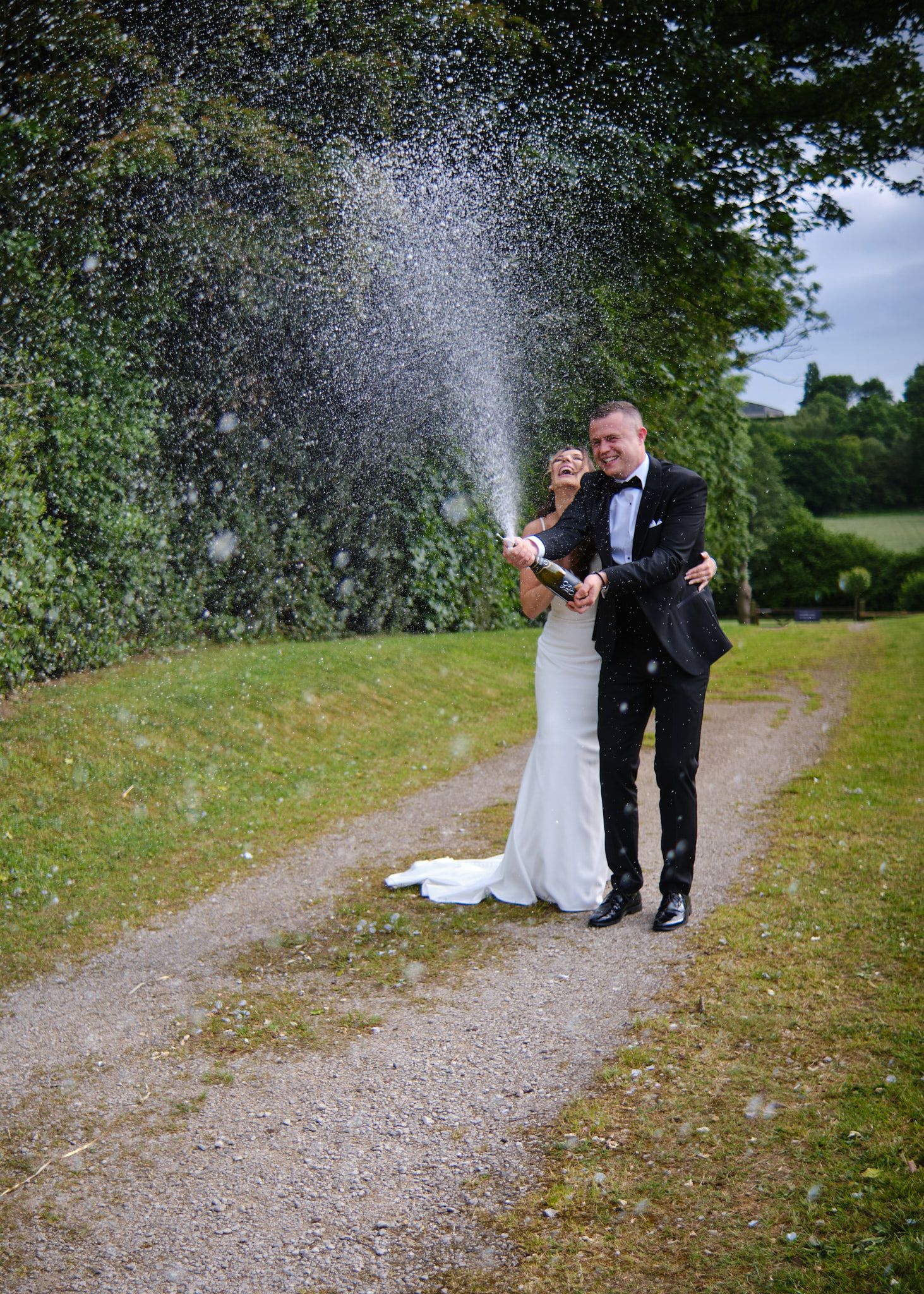 Bride and groom celebrating with a champagne spray on a garden path surrounded by lush greenery.