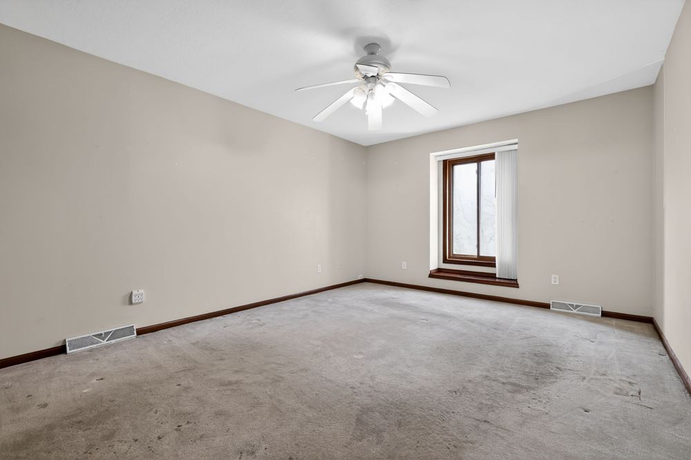 Empty room with beige carpet, neutral walls, ceiling fan, and a window with natural light and visible baseboard heating.