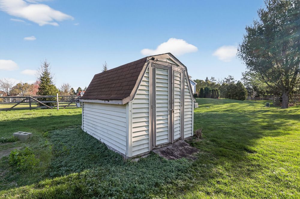 White gambrel-roofed storage shed on lush green lawn with trees and wooden fence in background on sunny day.