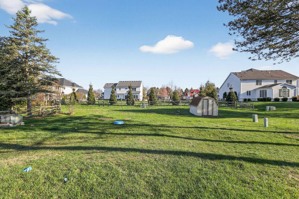 Spacious backyard with lush green lawn, small sheds, mature trees, and neighboring homes under a bright blue sky.