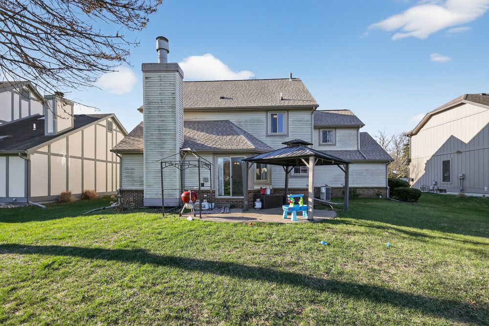 Suburban two-story home with chimney, gazebo, and spacious green backyard under a bright blue sky.