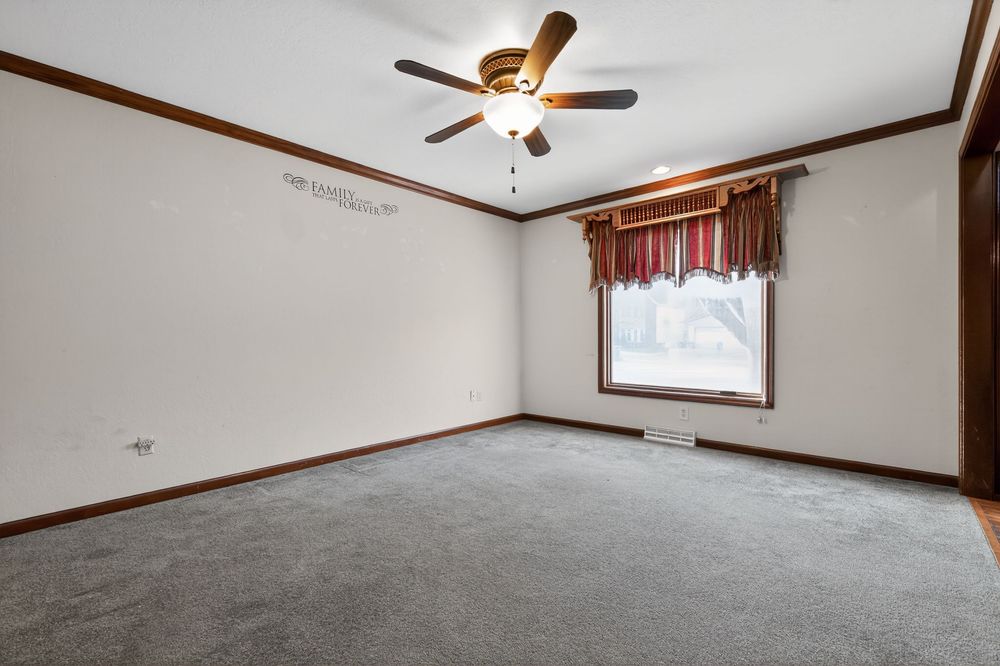 Empty bedroom with gray carpet, crown molding, ceiling fan, and a window with red curtains.