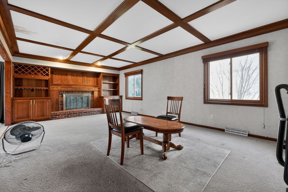 Spacious living room with coffered ceiling, built-in shelving, fireplace, and gray carpet.