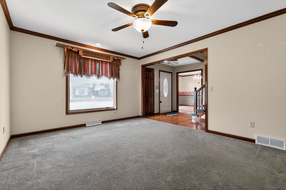 Spacious living room with gray carpet, crown molding, ceiling fan, and an open doorway leading to a hardwood foyer.