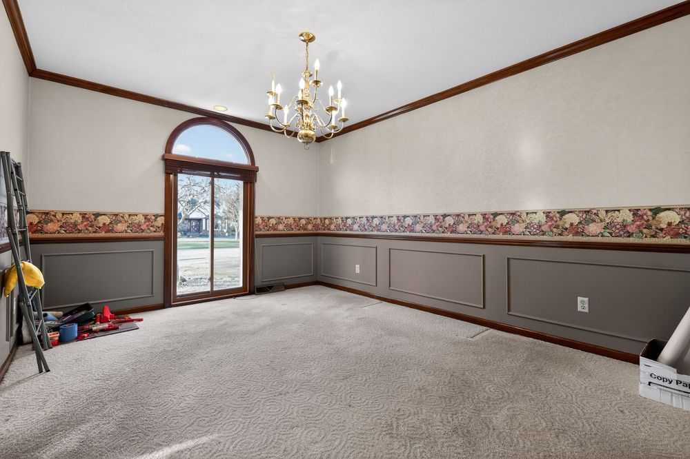 Elegant dining room with wainscoting, floral wallpaper border, chandelier, arched window, and neutral carpet flooring.