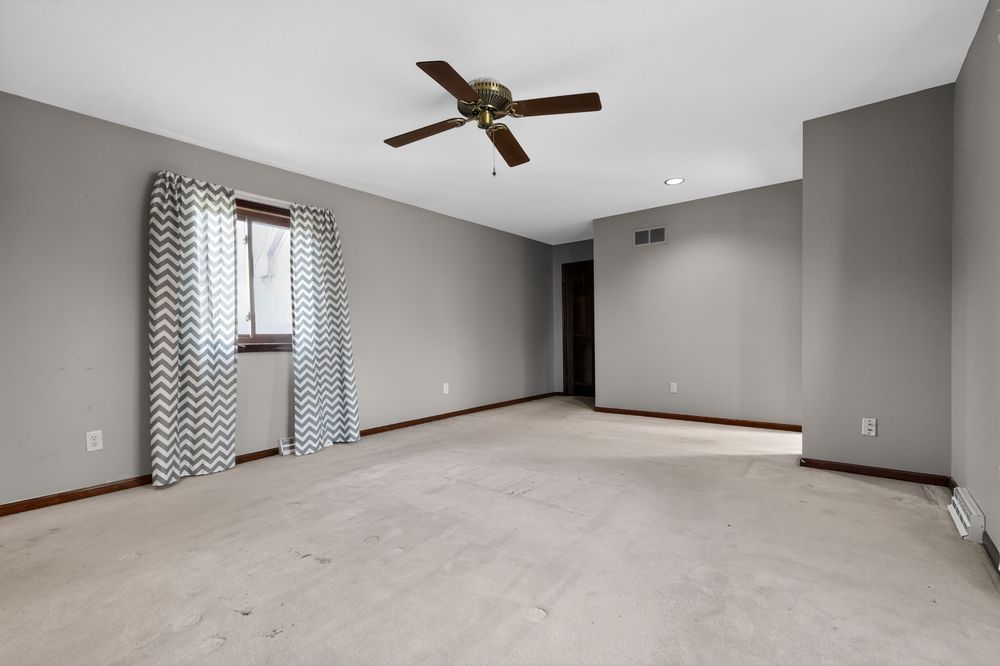 Spacious empty living room with gray walls, ceiling fan, chevron curtains, and neutral carpet flooring.