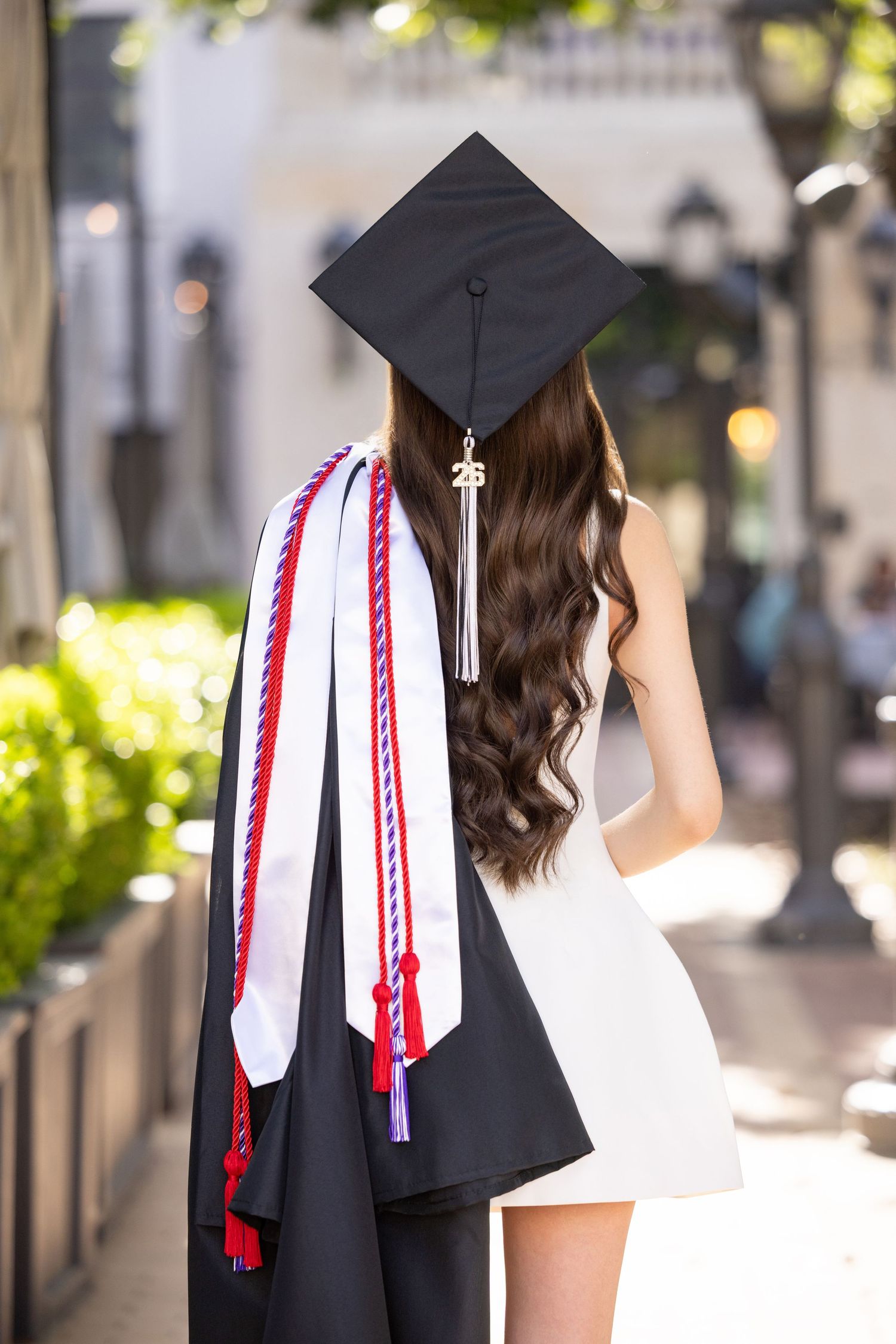 Graduate in black cap and gown holding honor cords, showing long wavy hair, standing outdoors on a sunny day.