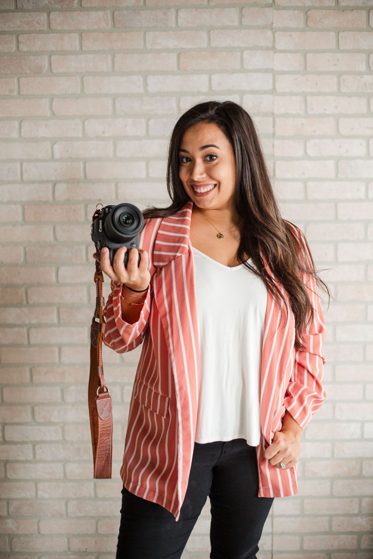 Smiling woman holding a DSLR camera wearing a red striped blazer and white top against a brick wall.