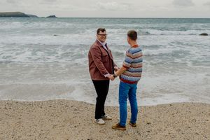 Two men hold hands romantically on a sandy beach with ocean waves crashing behind them.