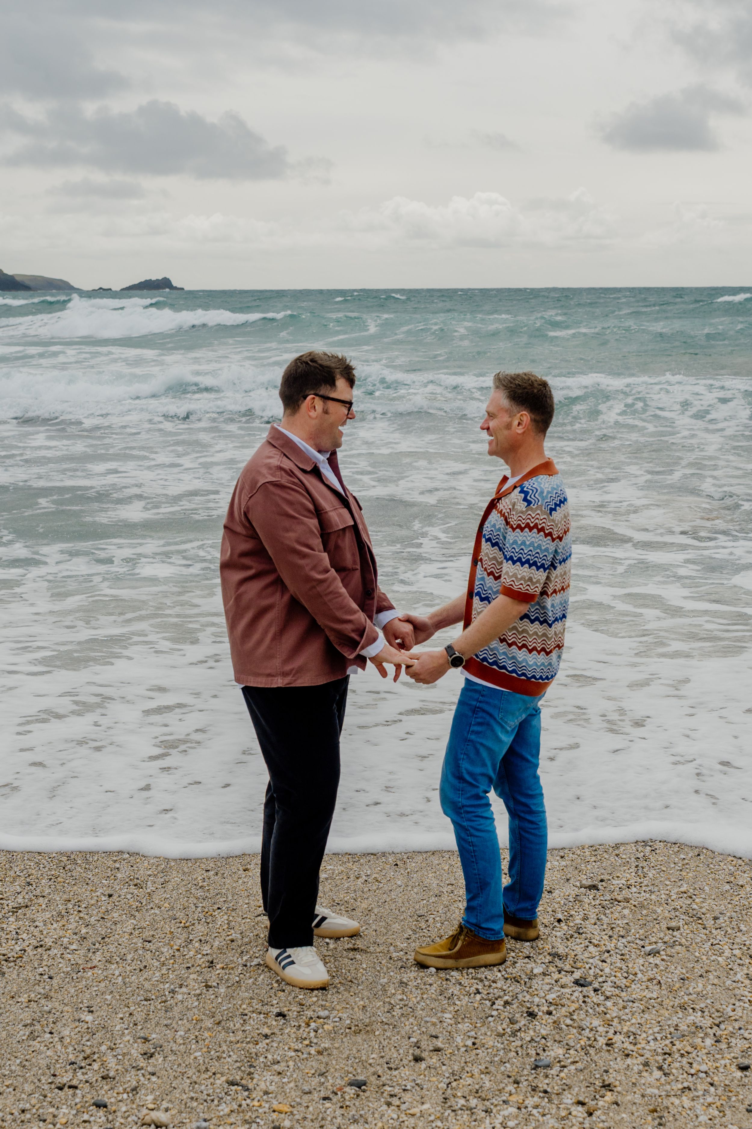 Two men holding hands on a pebble beach with ocean waves behind them on a cloudy day.
