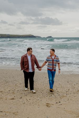 Two men holding hands and walking on a sandy beach with ocean waves and cloudy skies in the background.