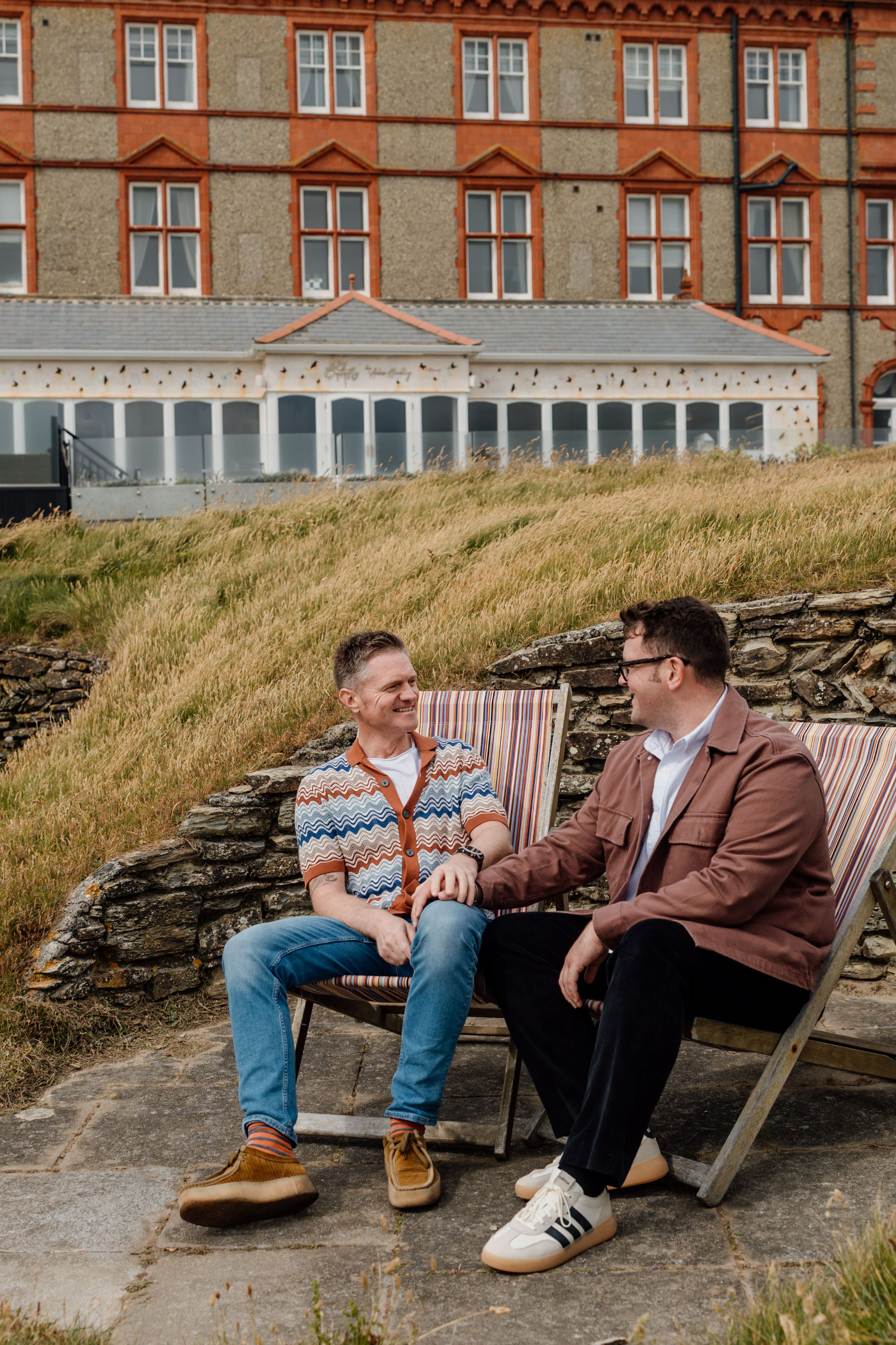 Two men sit in striped deckchairs chatting outside a grand stone building with manicured grounds.