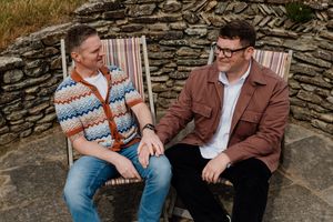 Two men sit in striped deck chairs holding hands and smiling at each other against a rustic stone wall.