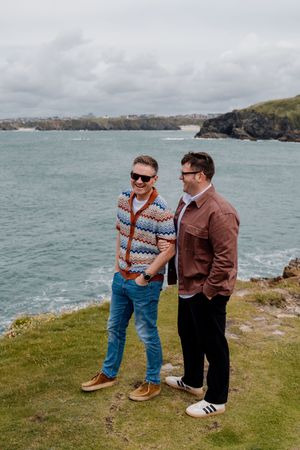 Two men laughing together on a coastal cliff, one in a colorful cardigan and jeans, the other in a brown jacket.