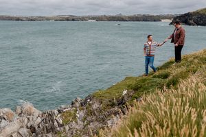 Two people stand on a rocky coastal cliff edge overlooking a scenic turquoise bay under a cloudy sky.