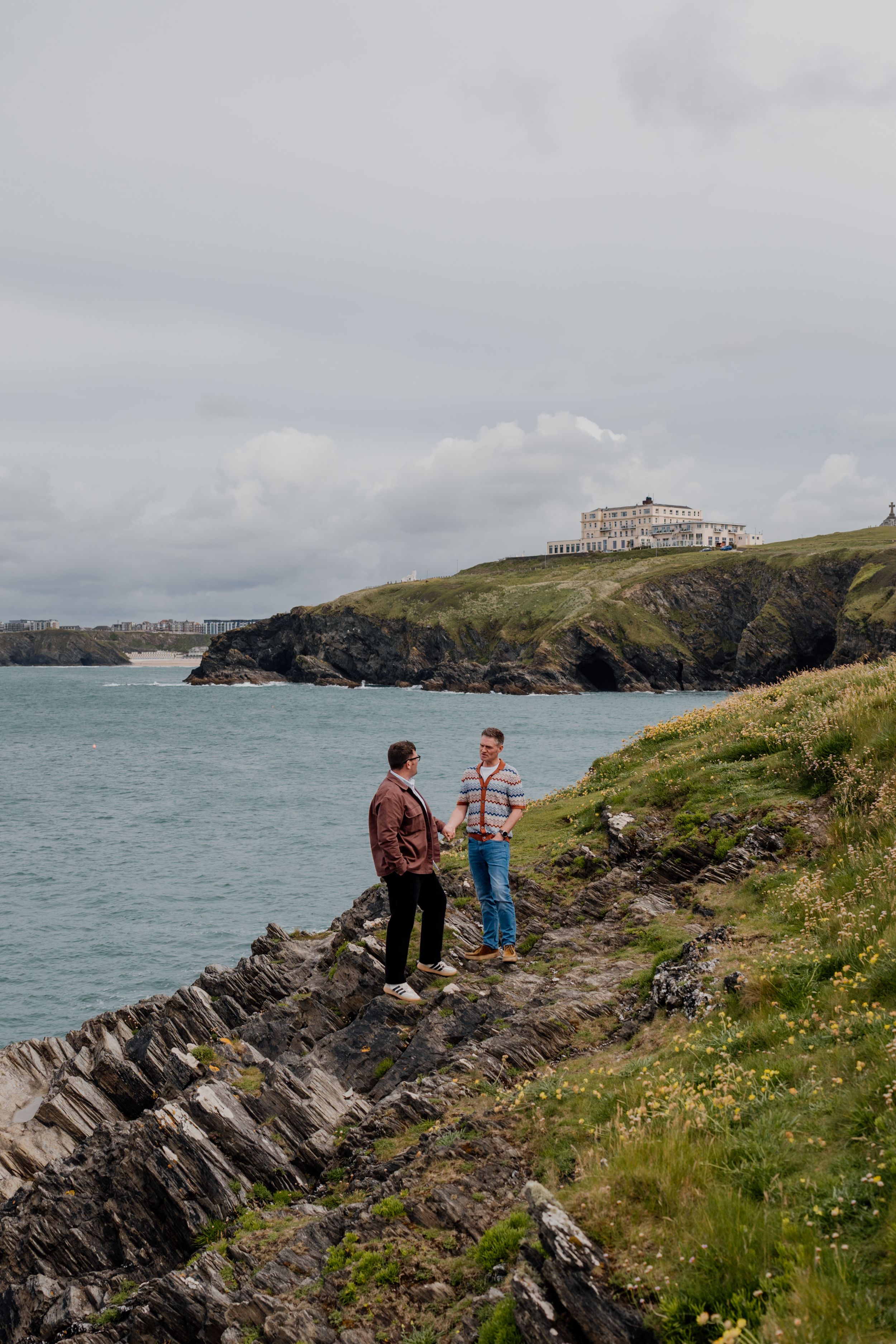 Two men stand on rocky coastal cliffs overlooking a turquoise bay with a grand building on the green hillside.