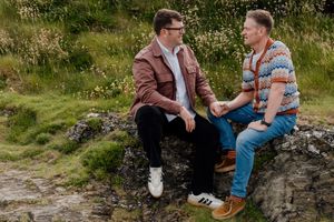 Two men sit on rocks in a lush green landscape, engaged in an intimate conversation outdoors.