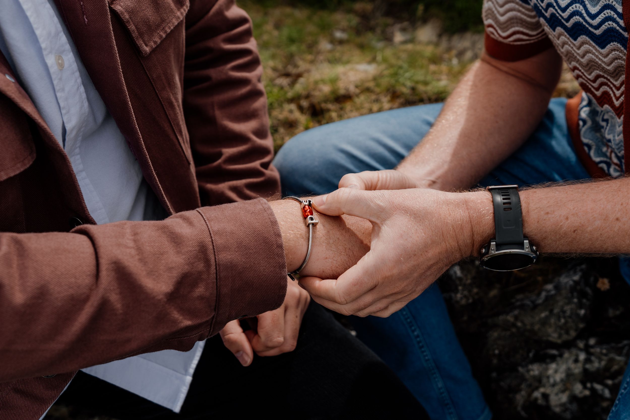 Two people sitting closely together holding hands, one wearing a red bracelet and brown jacket, the other a black watch.