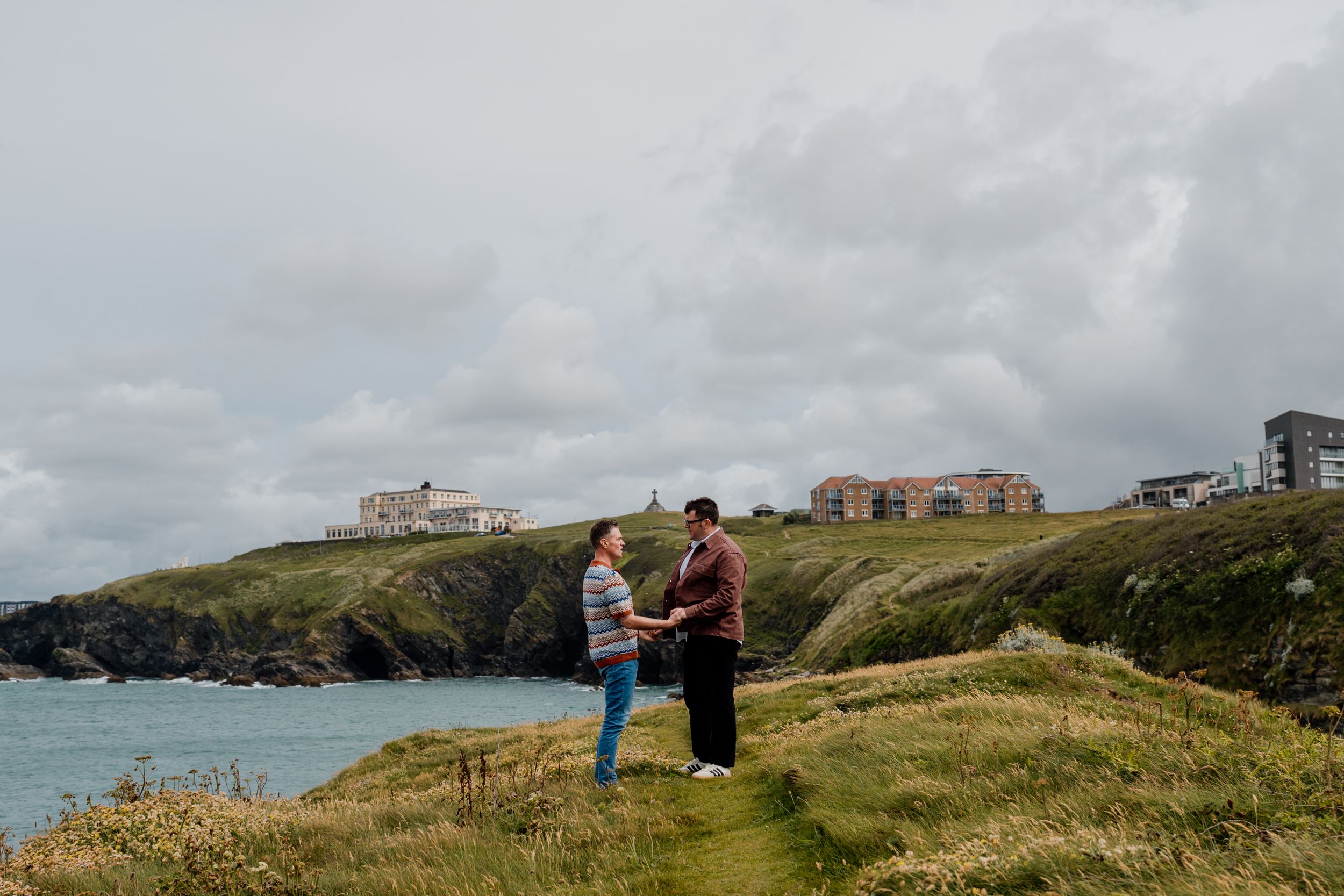Two people hold hands on a coastal cliff with buildings and ocean views under a cloudy sky.