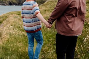 Couple holding hands walking along a scenic coastal grassy path, woman in colorful striped sweater and blue jeans.