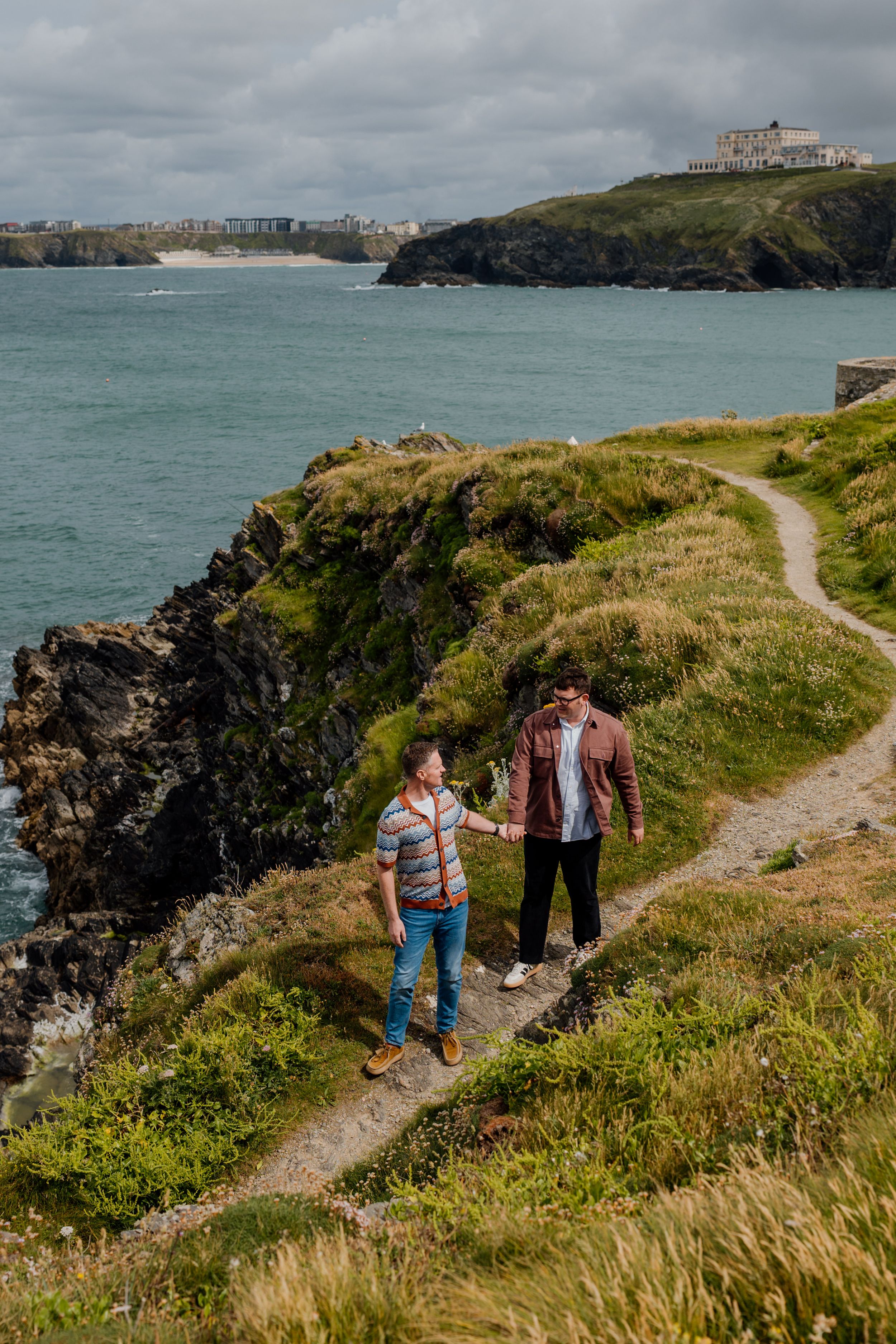 Two men hold hands while walking a scenic coastal cliff path with turquoise sea views.