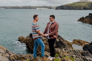 Two men holding hands on rocky coastal cliffs with turquoise ocean waves and green headland in background.