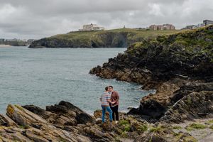 Couple standing on rocky coastal cliffs overlooking a turquoise bay with green headlands under cloudy skies.