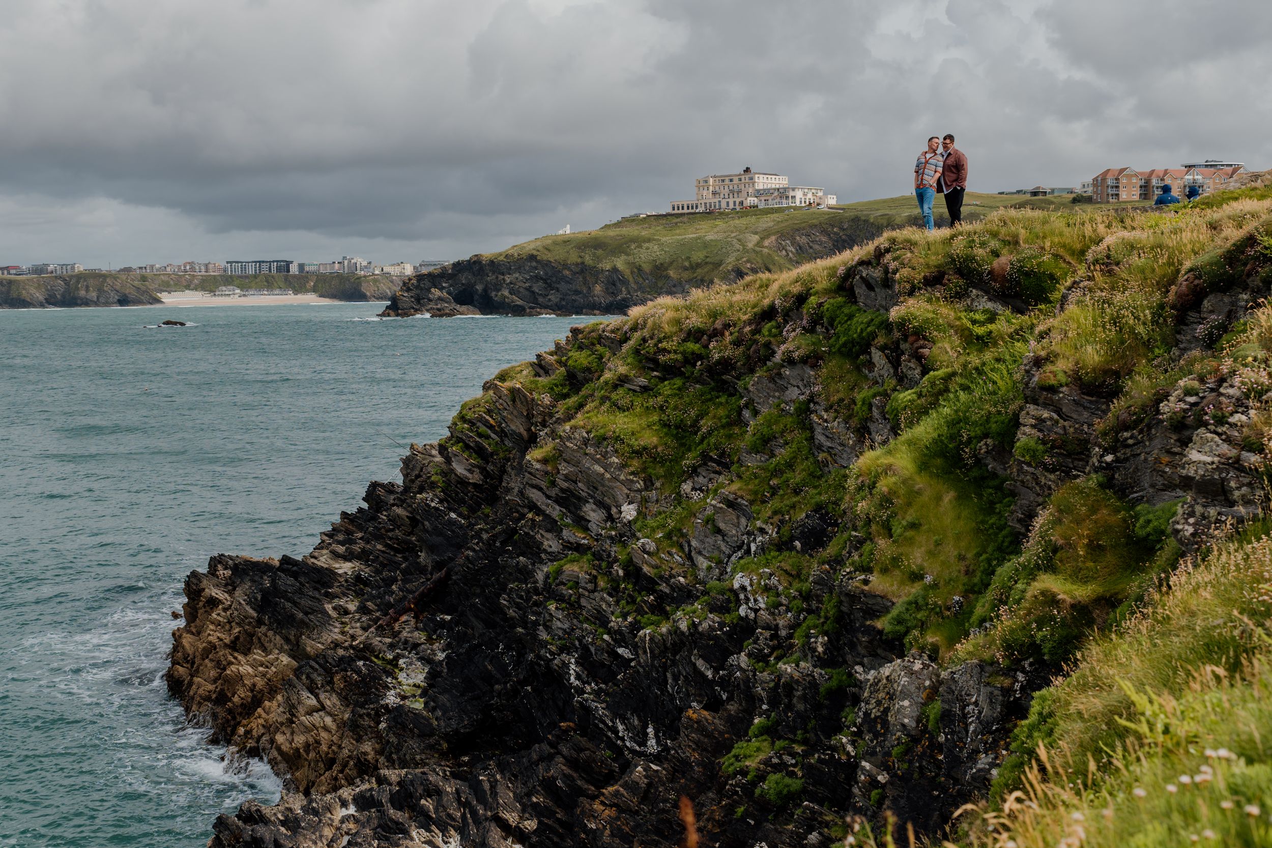 Couple standing on dramatic green coastal cliffs overlooking turquoise sea with a cloudy sky and distant town.