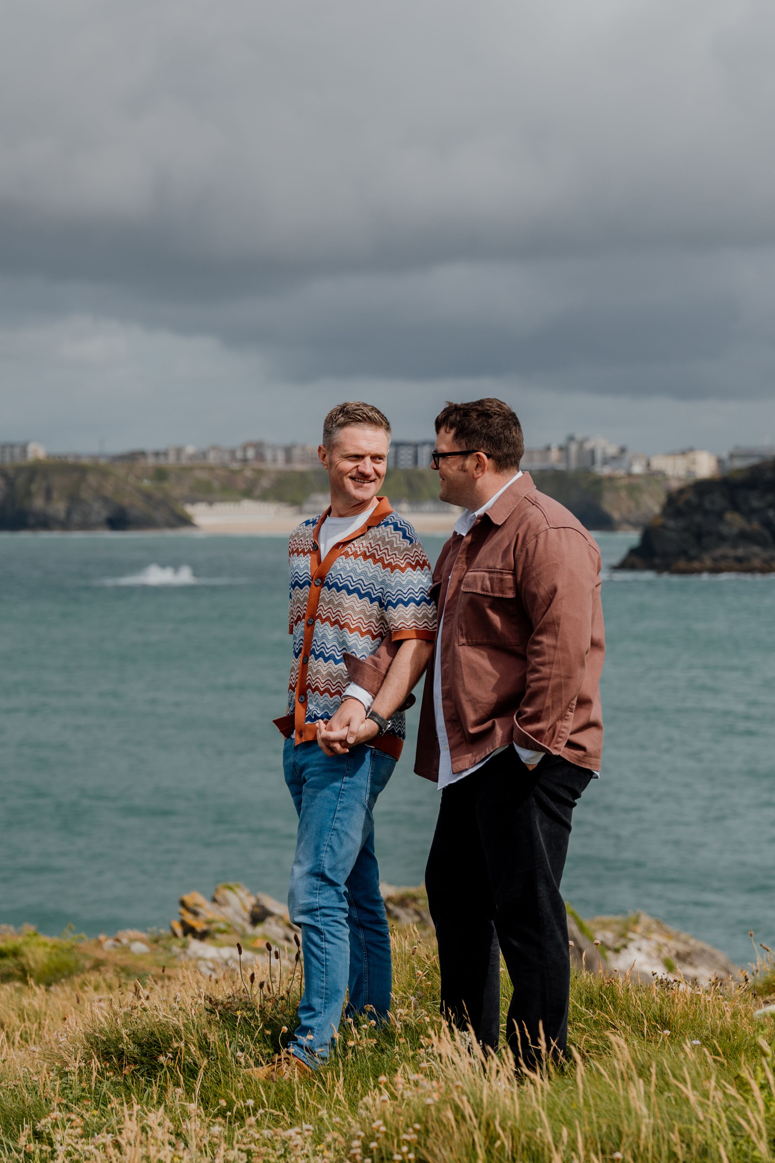 Two men hold hands smiling on a coastal cliff with turquoise sea and town in background.