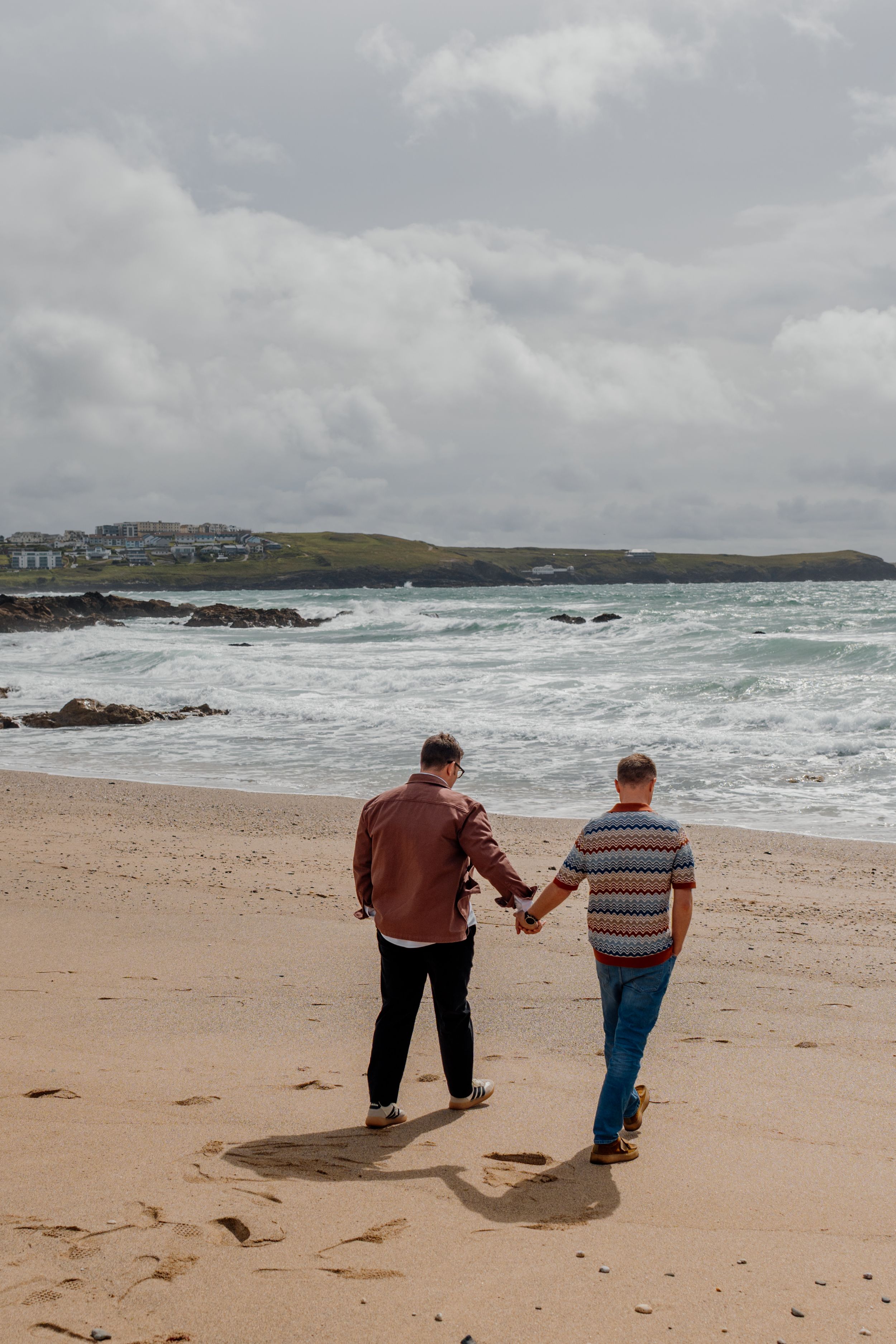 Two men holding hands walking along a sandy beach with ocean waves and cloudy skies in the background.