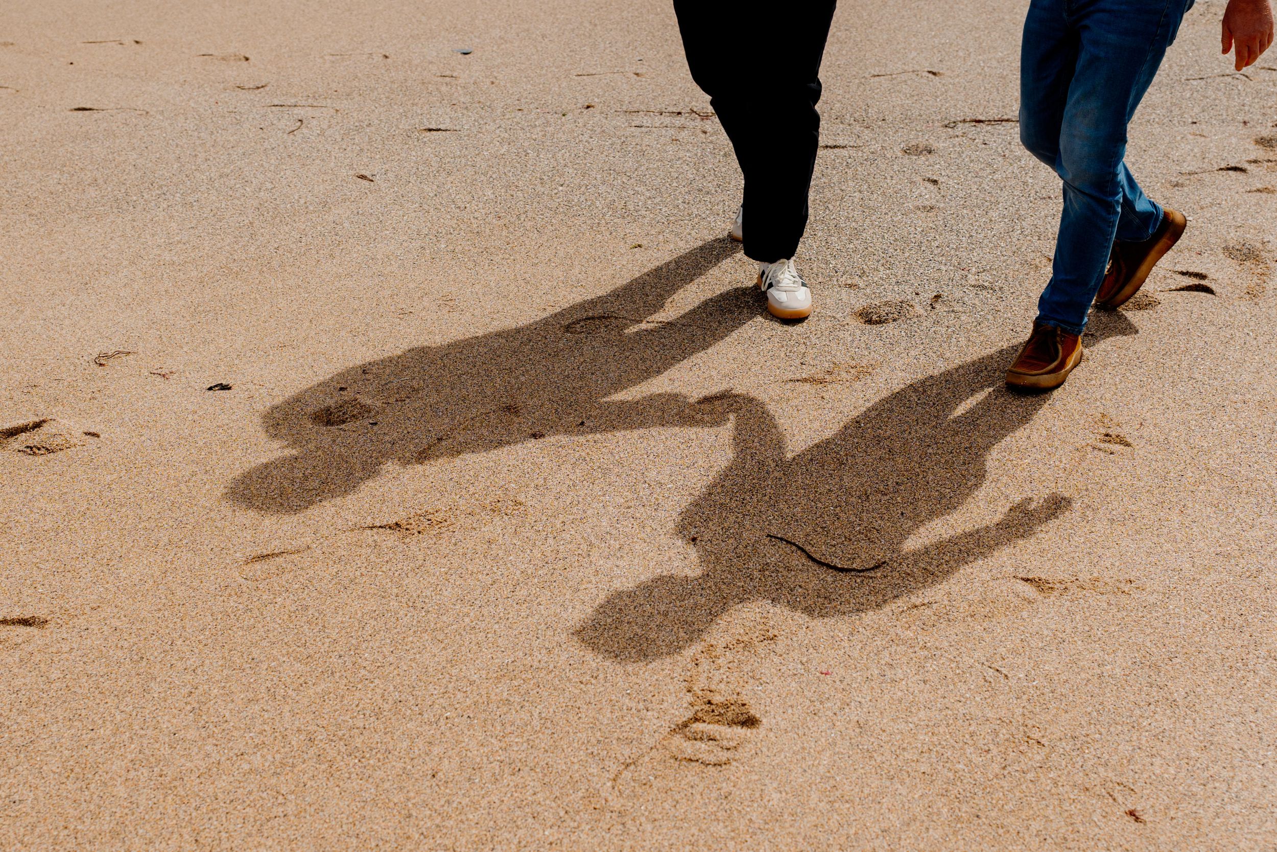 Shadows of two people walking together cast on sandy beach in bright sunlight.