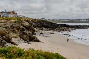 A lone person walks along a sandy beach beside rocky cliffs with white coastal buildings under overcast skies.