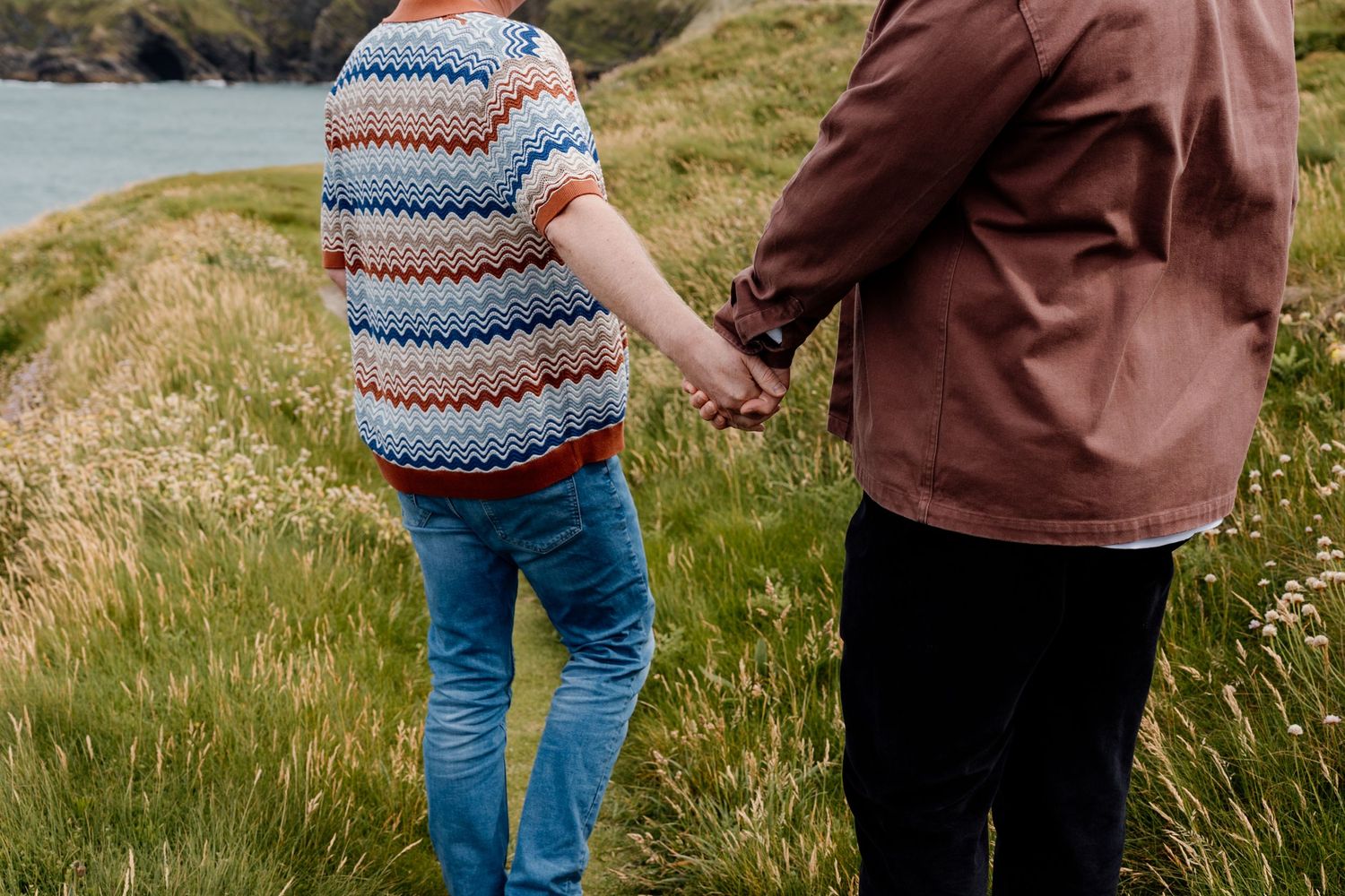 Couple holding hands walking along a scenic coastal grassy path, woman in colorful zigzag sweater and blue jeans.