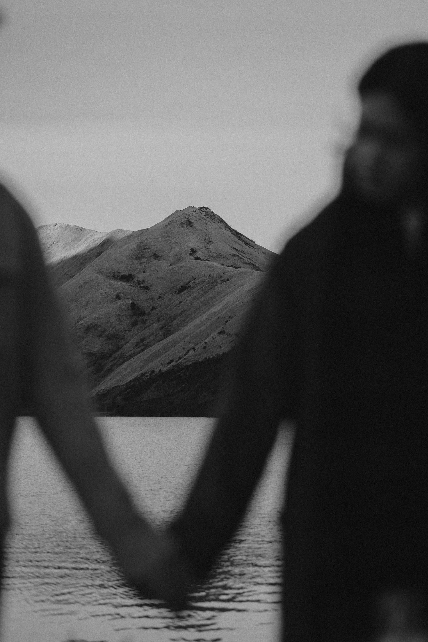 Black and white photo of a smiling couple holding hands with snow-capped mountains and a lake in the background.