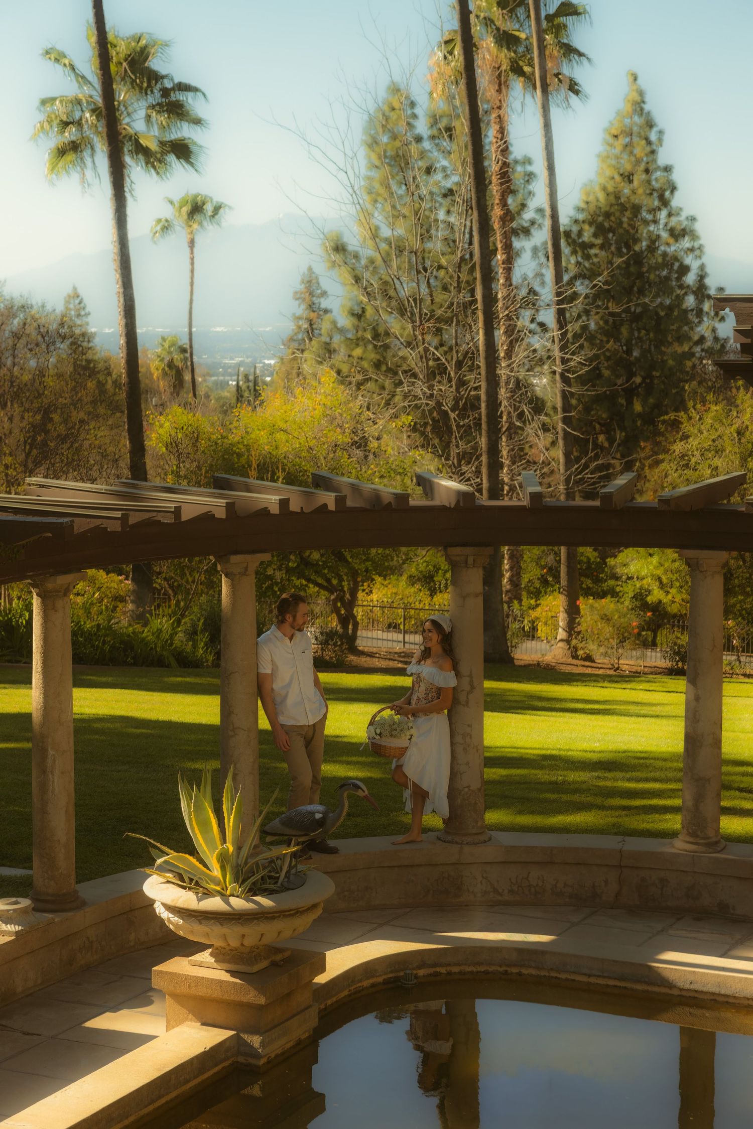 Couple standing under a pergola by a reflecting pool in a lush garden with palm trees on the grounds of the Kimberly Crest House & Gardens.