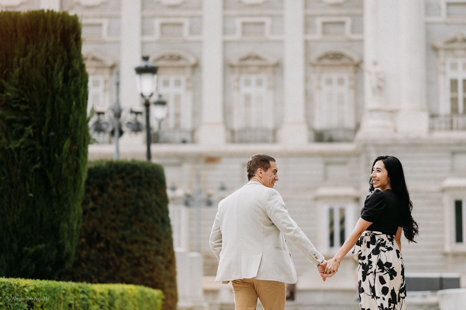 Couple holding hands walking away toward the Royal Palace of Madrid gardens.