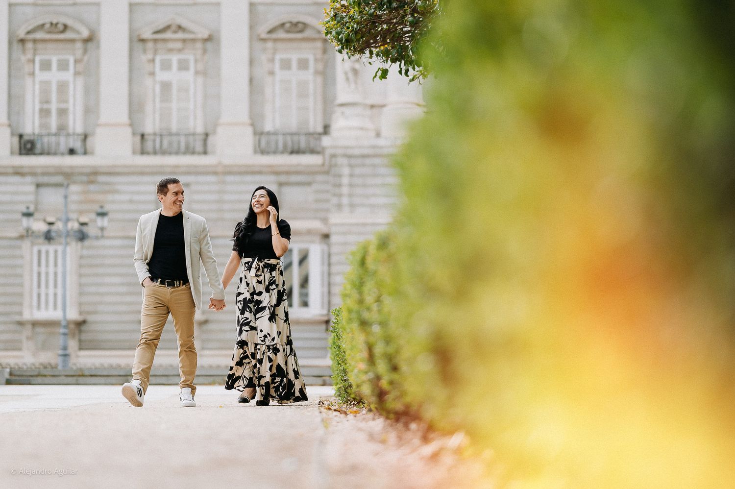 Couple walking hand in hand along a garden path near a grand white palace.