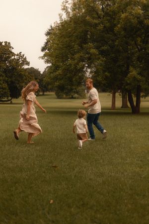 Family playing together in a lush green park, parents chasing their toddler on a cloudy day.