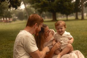 Happy young family sitting on grass in park, parents laughing with red-haired baby boy on sunny day.