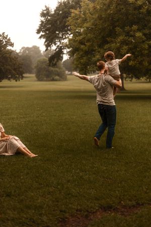 Father carrying child on shoulders with arms outstretched in green park while mother relaxes on grass.