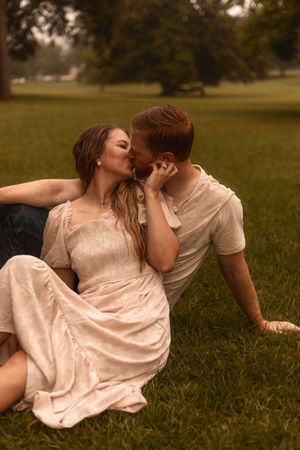Romantic couple sharing a kiss while sitting on green grass in a park during golden hour.