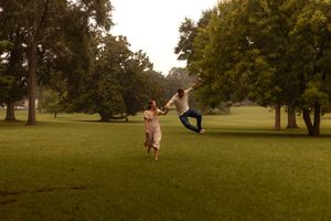 Couple dancing joyfully in lush green park, man leaping while holding womans hand.