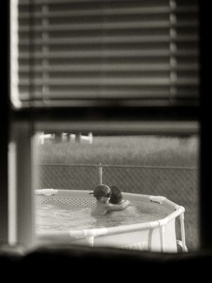 Black and white photo of two people relaxing in an above-ground pool, viewed through a window with blinds.