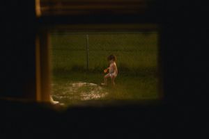 Child playing in backyard sprinkler at dusk, viewed through a window frame with green grass.