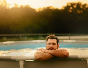 Man relaxing in an above-ground pool at golden sunset with lush green trees in the background.