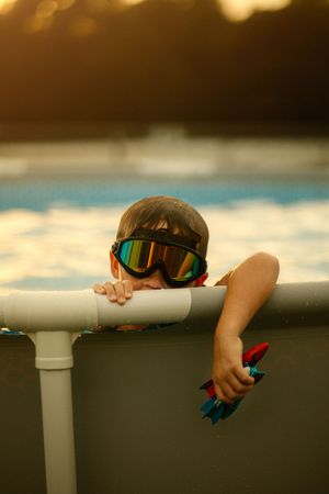 Young child wearing colorful ski goggles peeks over pool edge at sunset, holding a toy.