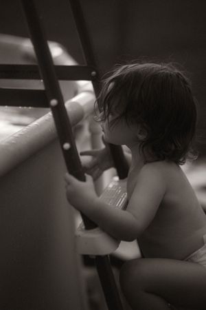 Black and white photo of a young toddler in a diaper climbing a ladder near a pool.